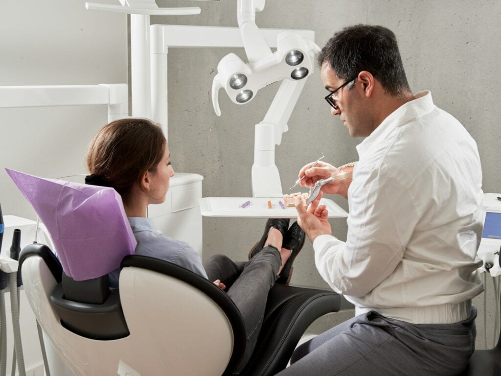 Male dentist in glasses showing a dental jaw model to a female patient sitting in a dental chair